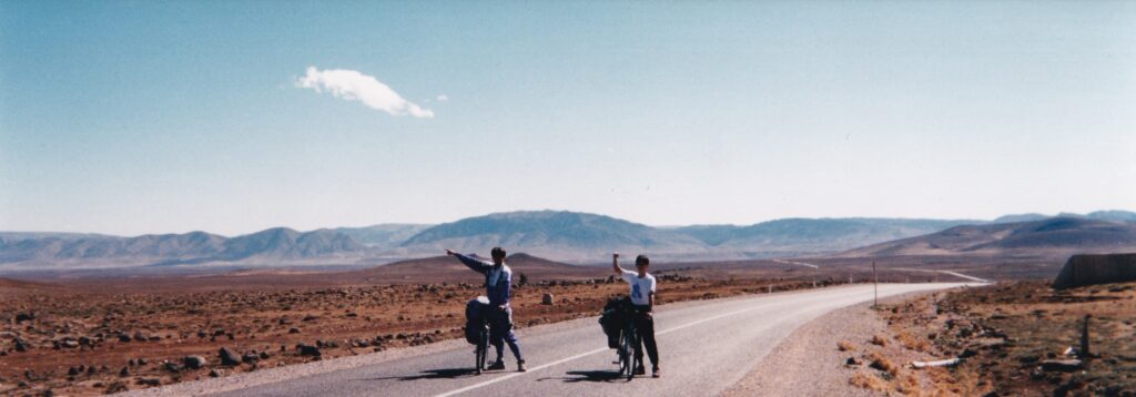 モロッコの砂漠地帯を、自転車に荷物を積んで走る旅人たち。荒涼とした大地と遠くの山並みが広がる中、仲間同士が手を挙げて進む風景。