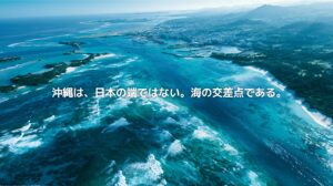 沖縄本島の海岸線、エメラルドブルーの海、サンゴ礁、都市部、港、遠くに連なる島影を俯瞰で捉えた風景。沖縄が日本の端ではなく、海の交差点であることを表現したアイキャッチ画像。