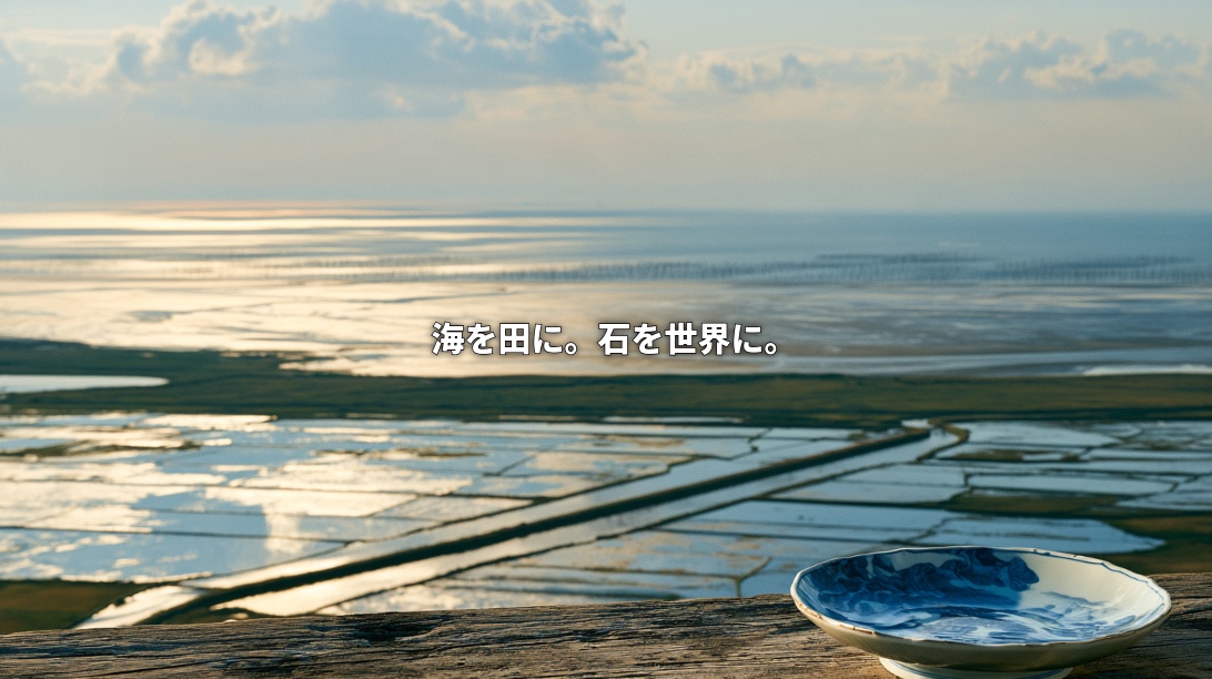 Illustration of rice fields and irrigation creeks stretching toward the Ariake Sea in Saga, with a blue-and-white Arita porcelain dish in the foreground and Japanese text reading “Turning sea into farmland, bringing stone to the world.”